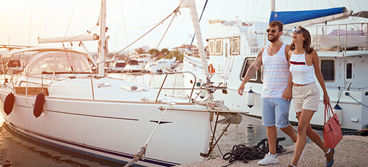A man and a woman in summer clothes walk along a dock with boats in the background